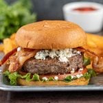 A close-up of a bacon blue cheese burger with a beef patty, crispy bacon strips, crumbled blue cheese, lettuce, and tomato on a toasted bun, served on a metal tray with fries and a cup of ketchup in the background.