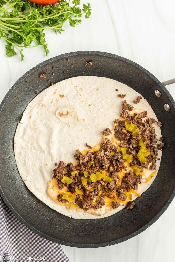 Large tortilla in a skillet topped with sauce, ground beef and diced pickles.