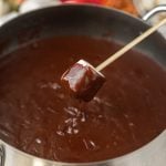 A marshmallow on a skewer is being dipped into a pot of rich chocolate fondue.