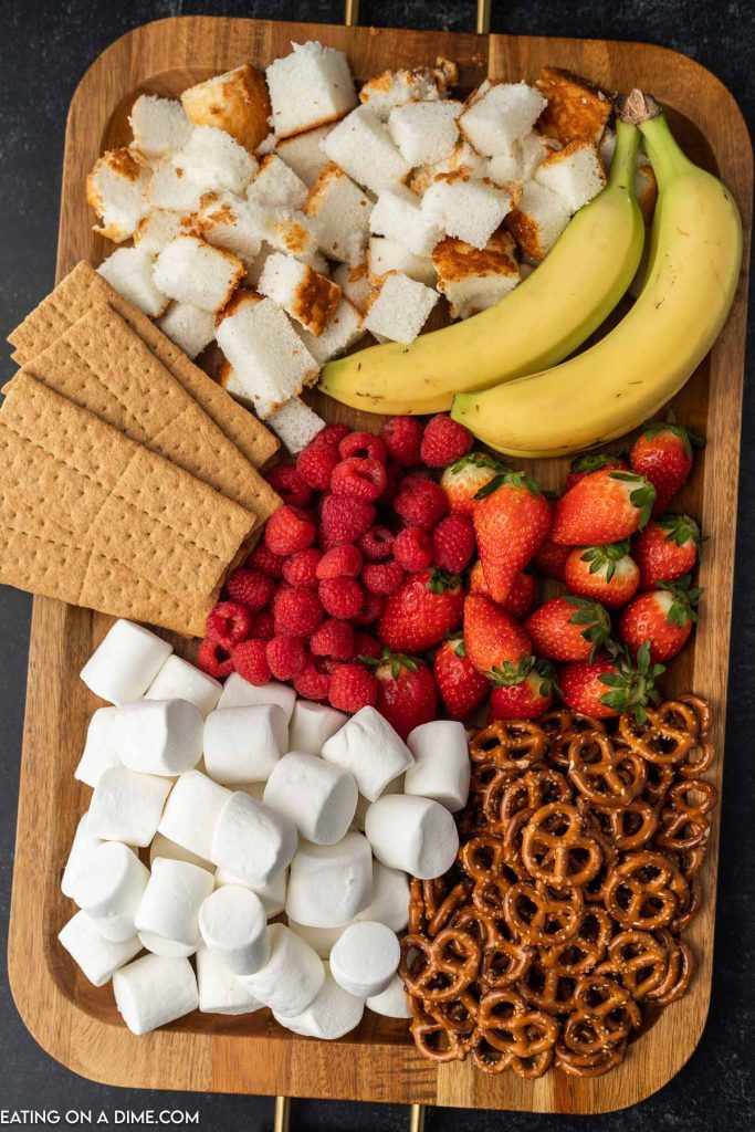 A platter of bananas, pound cake, graham crackers, raspberries, strawberries, marshmallows and pretzels