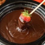 A fresh strawberry on a skewer is being dipped into a bowl of smooth, melted chocolate made in a Crock Pot Chocolate Fondue. The strawberry is partially coated, and the rich chocolate covers its pointed end.