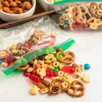 A clear ziplock bag with trail mix from a Trail Mix Bar—pretzels, Cheerios, almonds, colorful candies, raisins, and gummy bears—sits open with some mix spilled on a white surface. More trail mix and nuts are visible in containers behind.