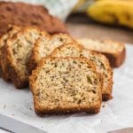 Several slices of Air Fryer Banana Bread are arranged on parchment paper, showcasing a moist, textured interior with visible banana and nut pieces. A whole loaf and a banana are blurred in the background.