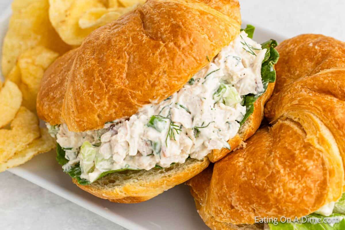 A flaky croissant is filled with a creamy chicken salad mixture, touted as the best chicken salad recipe, containing herbs and celery, garnished with fresh lettuce. The sandwich is served on a white plate alongside crispy potato chips.
