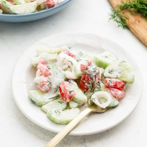 A fresh Creamy Cucumber Tomato Salad graces a white plate, featuring cucumber slices, tomato chunks, and onion rings draped in a creamy dressing. Garnished with dill sprigs and served with a fork, the scene is completed by a cutting board brimming with greens in the background.