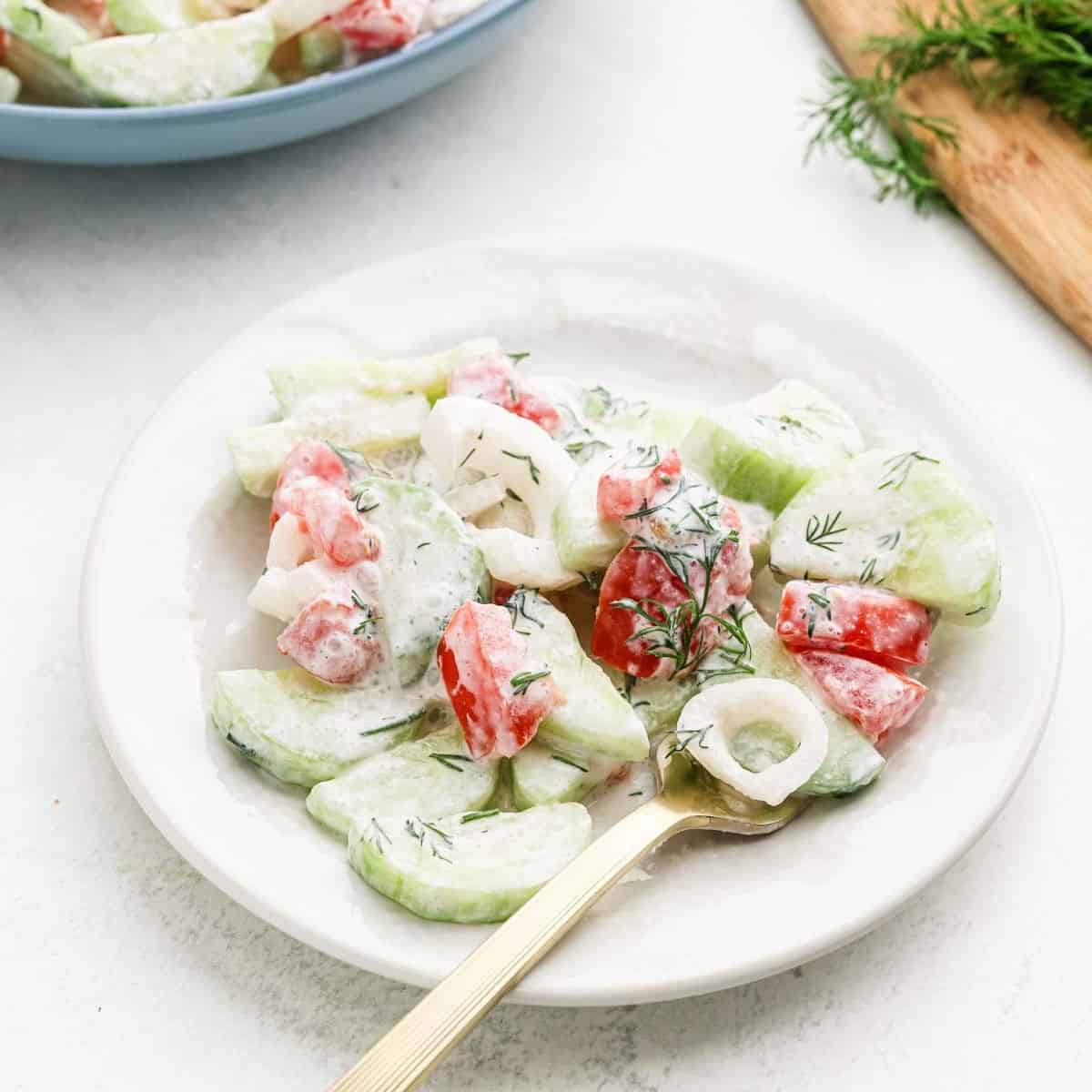 A fresh Creamy Cucumber Tomato Salad graces a white plate, featuring cucumber slices, tomato chunks, and onion rings draped in a creamy dressing. Garnished with dill sprigs and served with a fork, the scene is completed by a cutting board brimming with greens in the background.