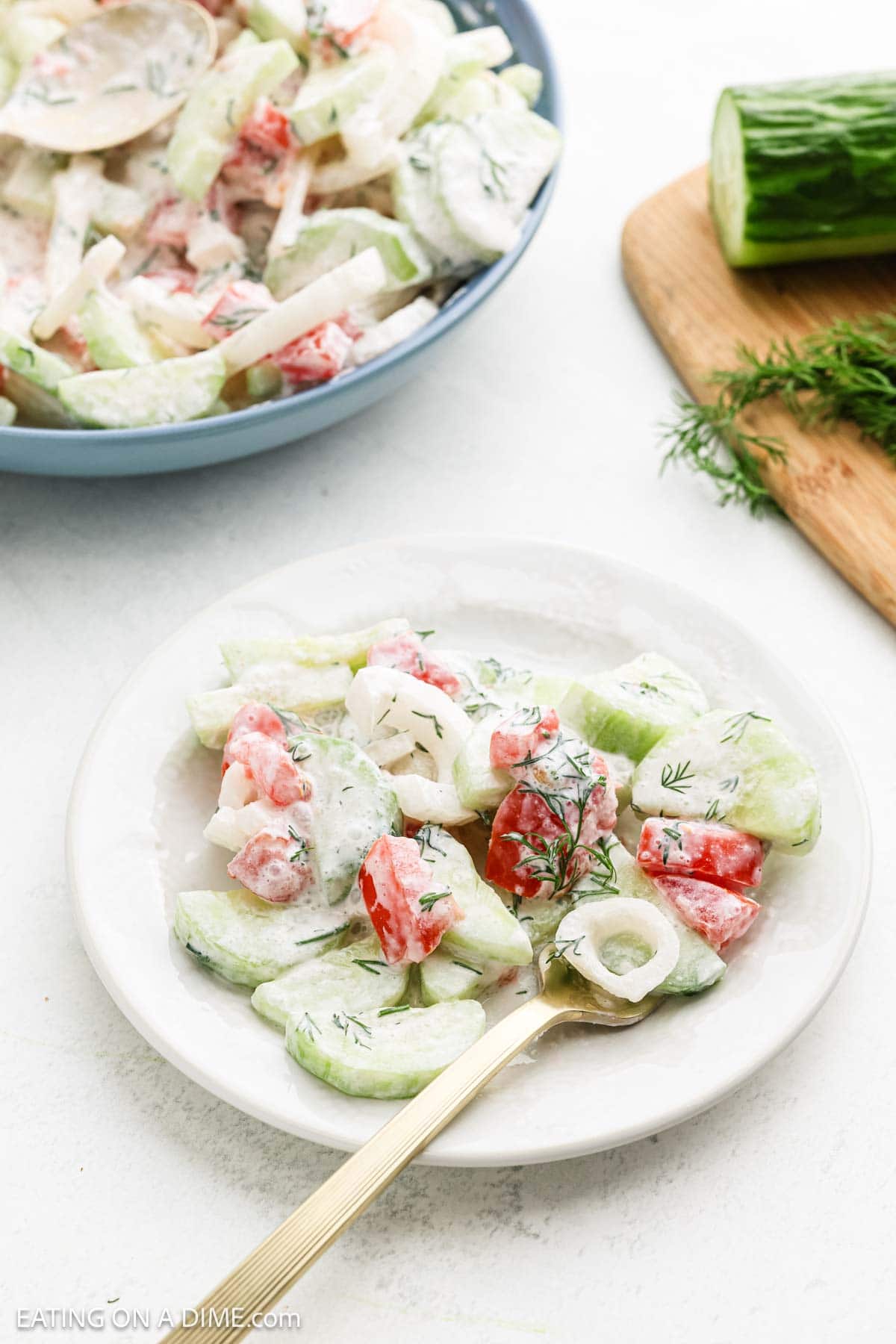 A plate of Creamy Cucumber Tomato Salad showcases sliced cucumbers, tomatoes, onions, and dill. A fork rests nearby. In the background, a larger bowl of this refreshing salad and a partial cucumber are seen on the countertop.