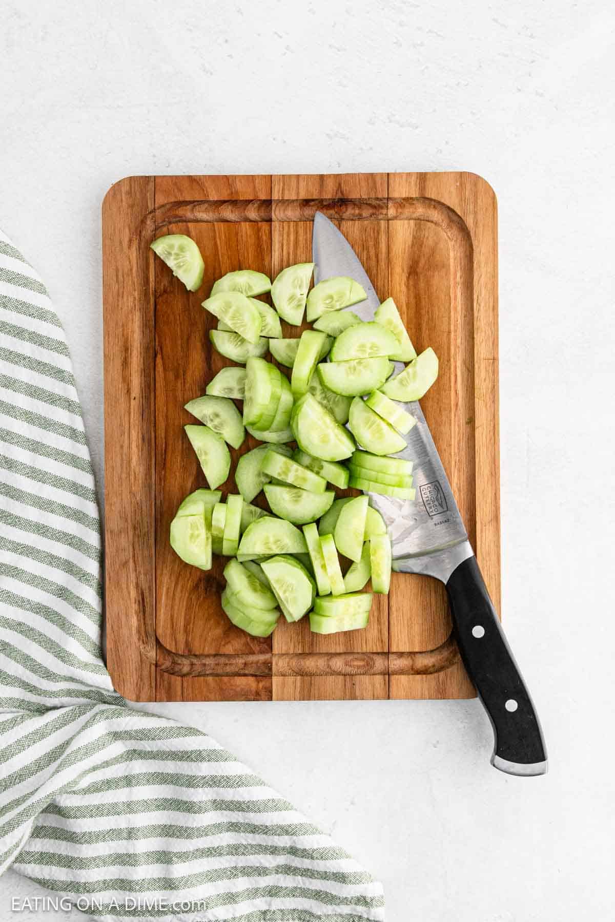 A wooden cutting board holds sliced cucumber pieces beside a large knife, ready for a refreshing Creamy Cucumber Tomato Salad. A striped kitchen towel is neatly placed beside the board, setting the perfect scene for culinary creativity.
