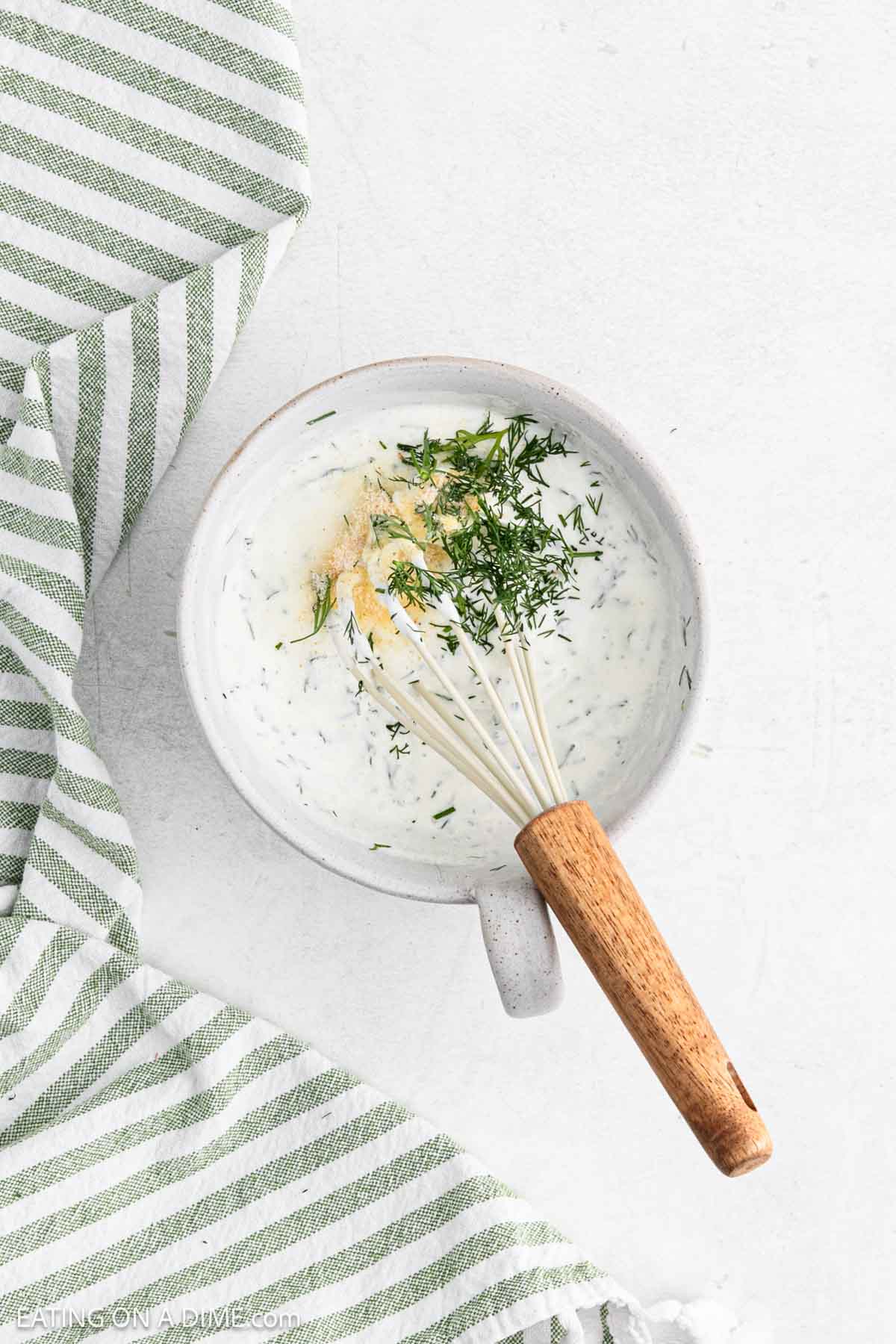 A ceramic bowl brimming with creamy white dill sauce, perfect for a refreshing Creamy Cucumber Tomato Salad, is topped with fresh dill and spices. A wooden-handled whisk rests inside, while a green and white striped cloth lies beside it on a light surface.