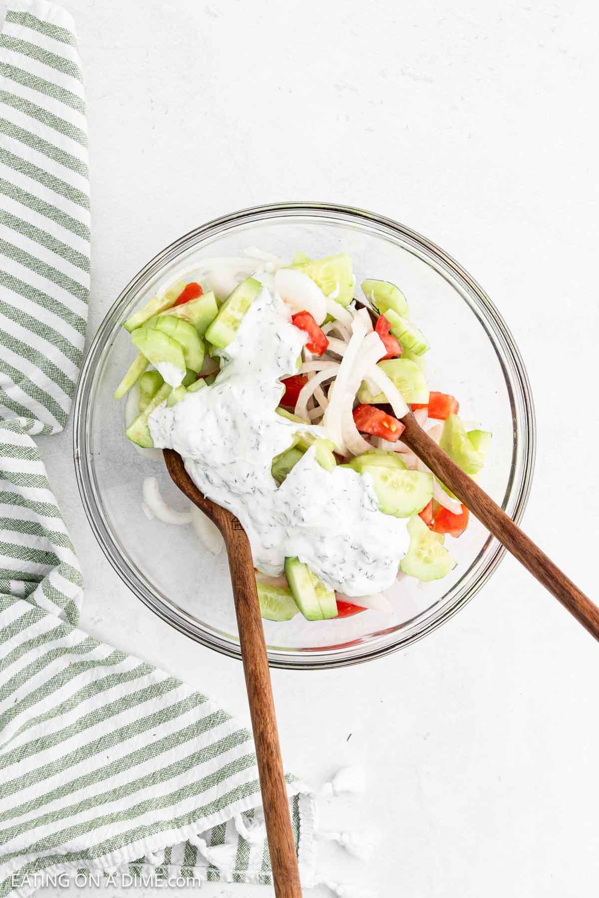 A glass bowl brimming with a Creamy Cucumber Tomato Salad is adorned with a luscious dressing. Inside, sliced cucumbers, tomatoes, and onions mix harmoniously. Two wooden spoons rest invitingly inside, while a green and white striped cloth lies beside the bowl.