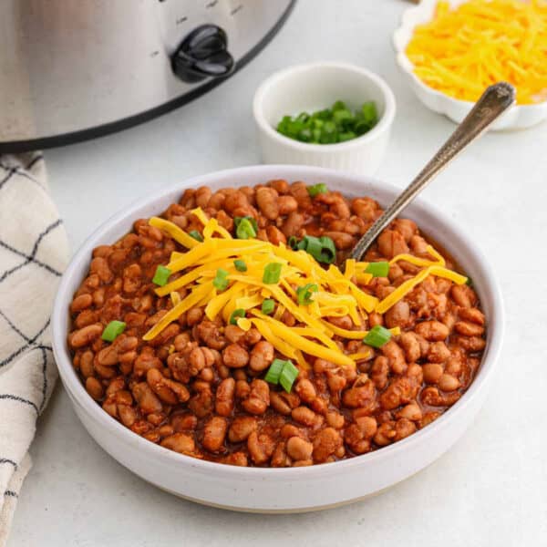 A bowl of crock pot ranch beans is topped with shredded cheddar cheese and sliced green onions. A spoon is placed inside. In the background, there's a small bowl of green onions, a plate of cheddar cheese, and a slow cooker. A checkered cloth is draped beside the bowl.