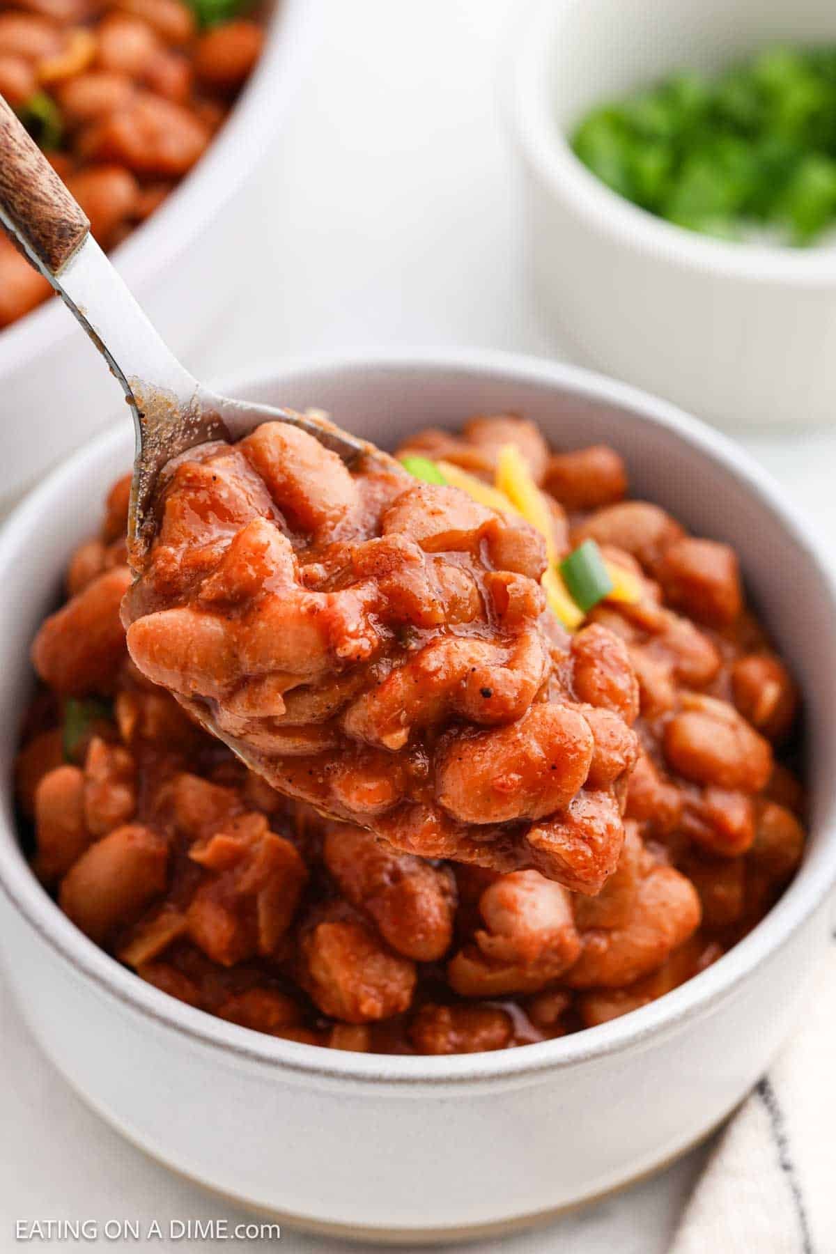 A spoonful of baked beans in a rich sauce is held above a white bowl filled with crock pot ranch beans. The background features a small dish with chopped green herbs.