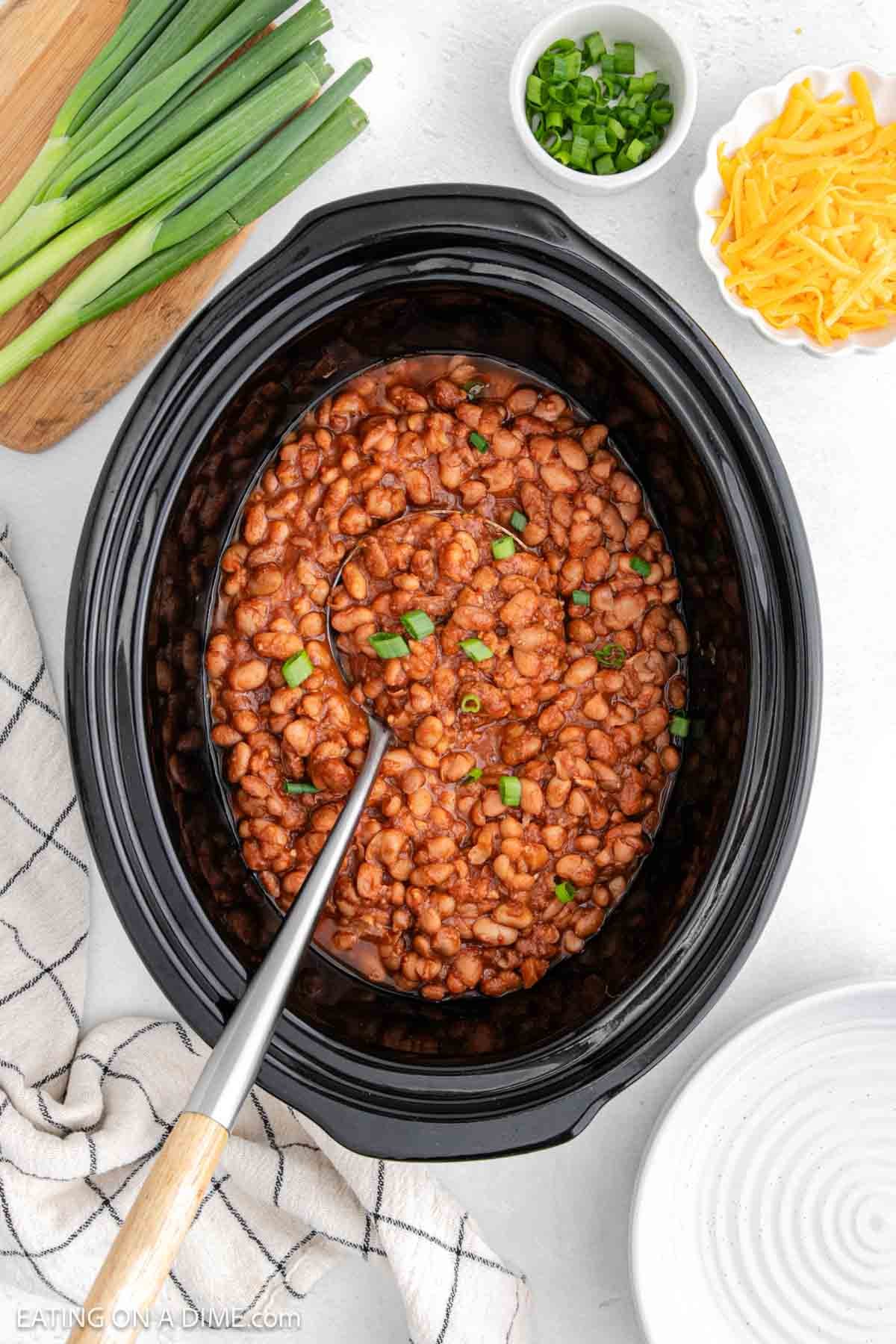 A black crock pot brimming with ranch-flavored beans, topped with chopped green onions, invites you to dig in with the ladle inside. Surrounding it are bowls of extra green onions and shredded cheese, a bunch of whole green onions, and a cozy kitchen towel.
