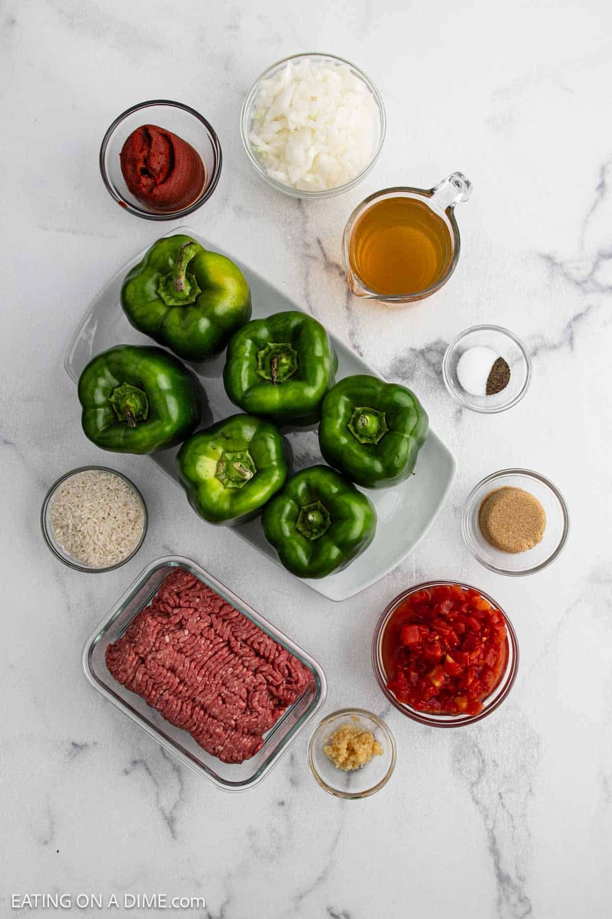 Top-down view of crock pot stuffed peppers recipe ingredients—green bell peppers, ground beef, diced onions, tomato paste, diced tomatoes, rice, garlic, broth, brown sugar, salt, and pepper—all arranged on a white marble surface.