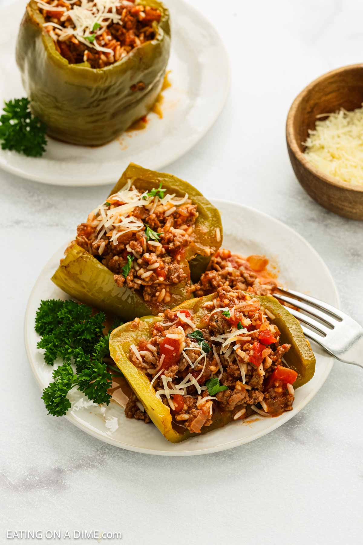 A plate with two green bell peppers stuffed with rice, ground meat, and tomato sauce—just like a classic crock pot stuffed peppers recipe—topped with shredded cheese and parsley, sits beside a fork. A whole stuffed pepper and cheese are in the background.