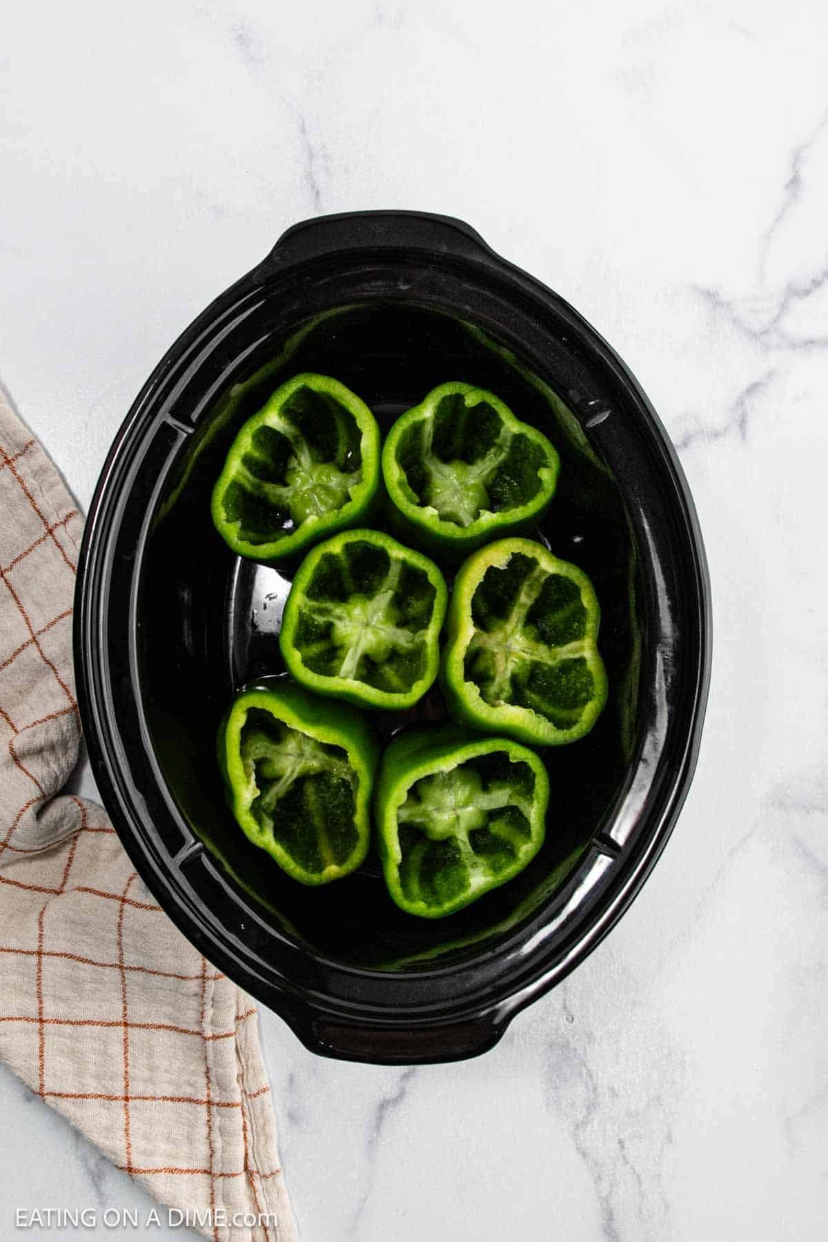 Six halved green bell peppers are arranged cut-side up in a black crock pot, ready for this delicious crock pot stuffed peppers recipe, set on a white marble surface with a beige and red plaid kitchen towel to the side.