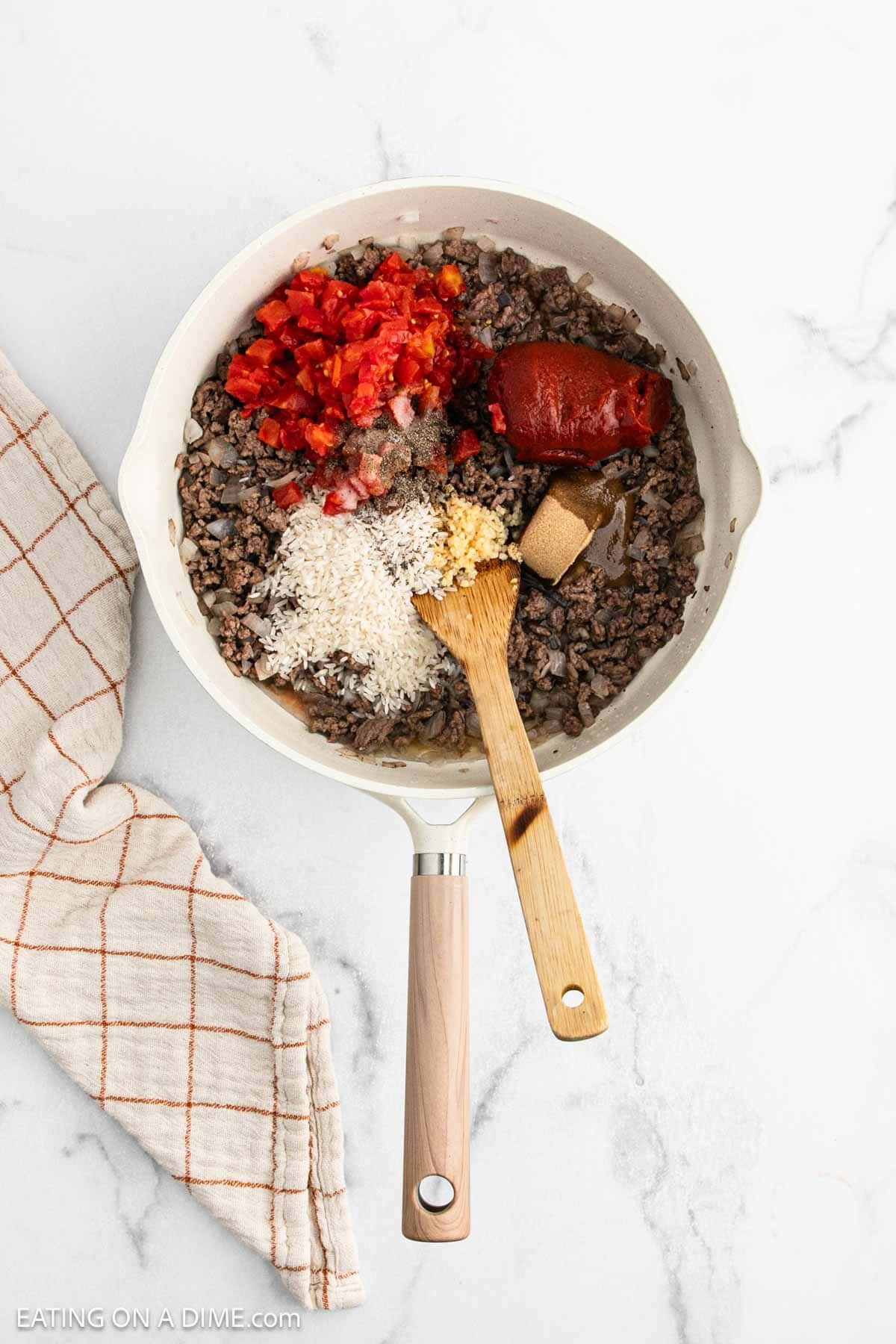 A skillet on a marble surface holds the savory filling for a crock pot stuffed peppers recipe—ground beef, chopped tomatoes, tomato paste, rice, onions, and spices—stirred with a wooden spoon. A cream and orange checkered towel rests beside the pan.