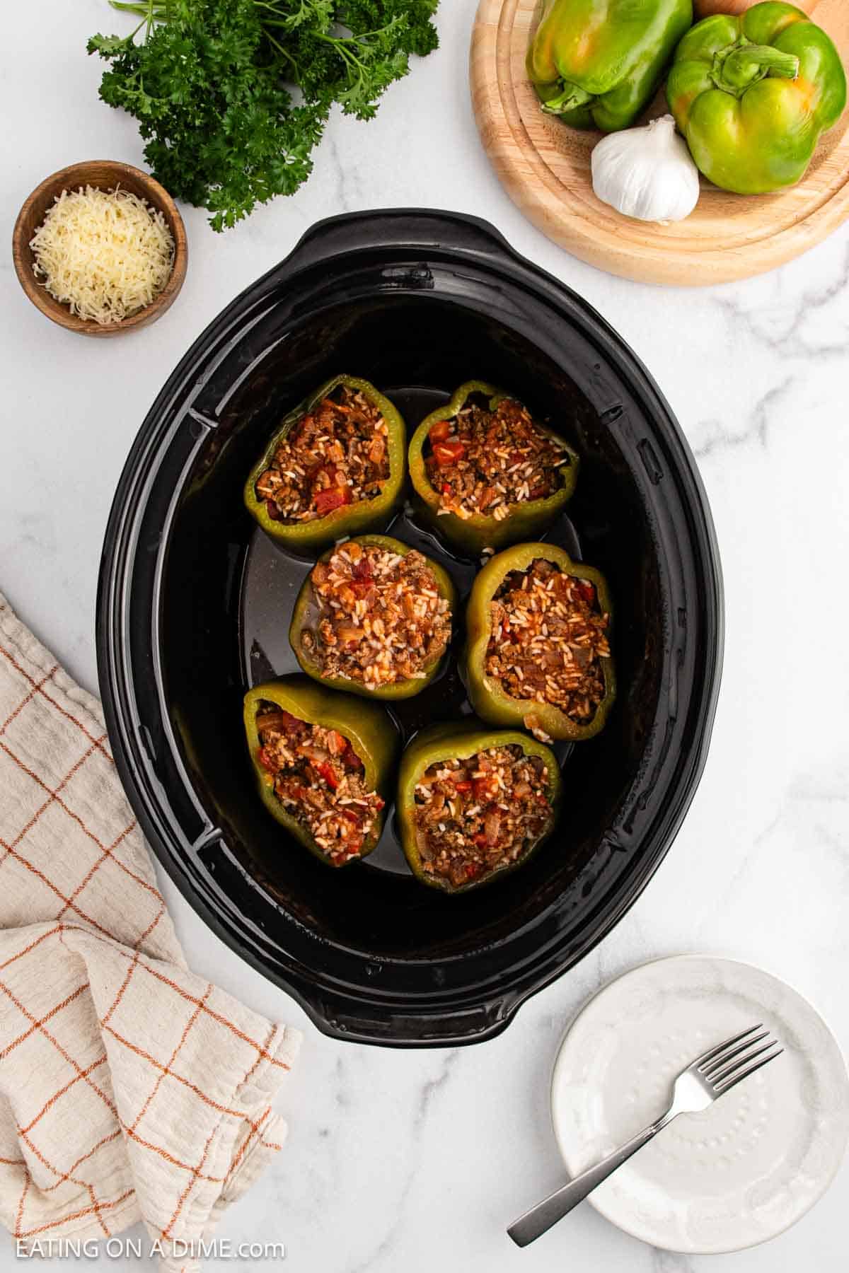 Overhead view of a crock pot stuffed peppers recipe: six green bell peppers filled with a savory meat and rice mixture. Surrounding the cooker are bell peppers, garlic, parsley, grated cheese, a napkin, and an empty plate with a fork.