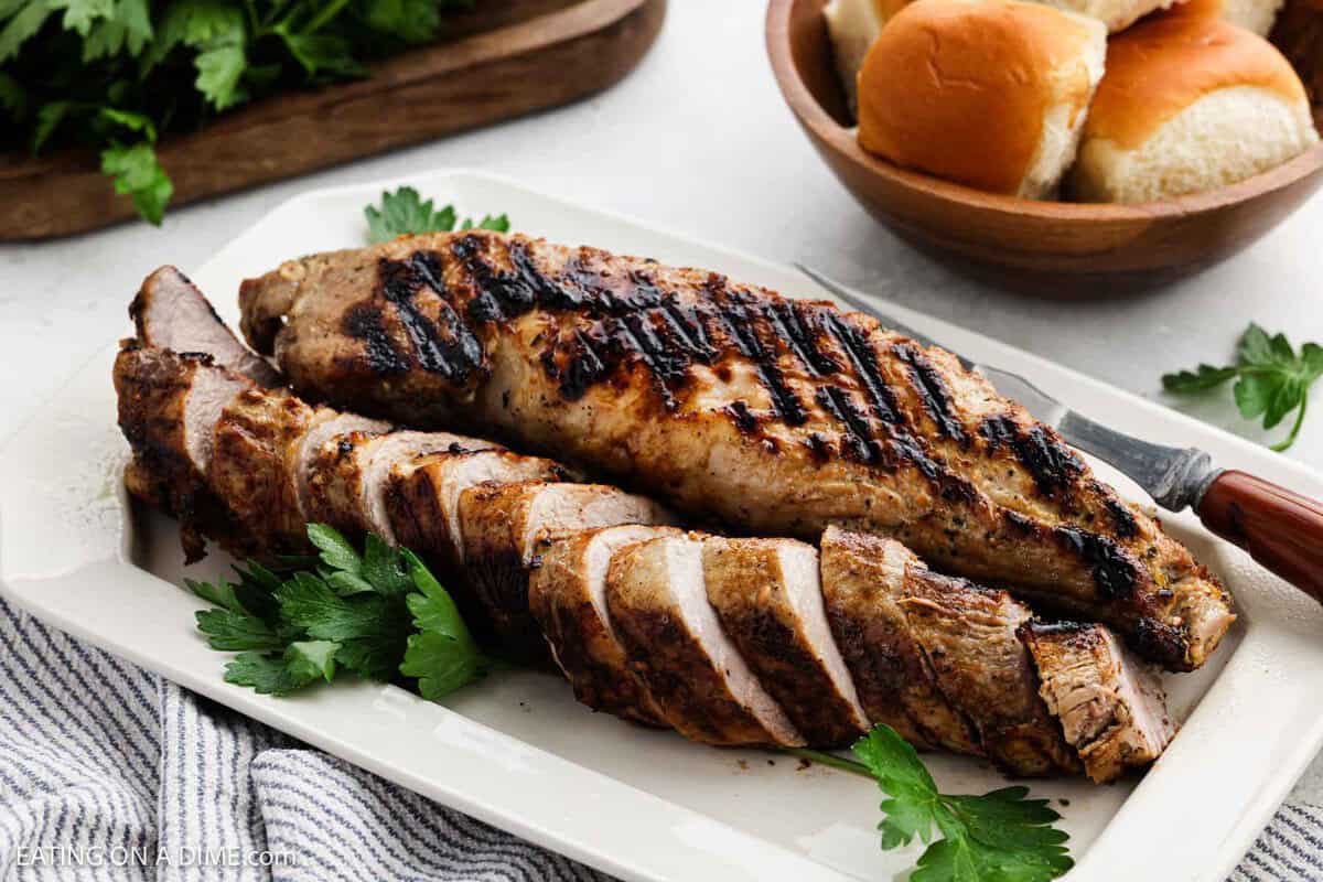 Sliced Grilled Pork Tenderloin on a white platter with a bowl of dinner rolls in the background.