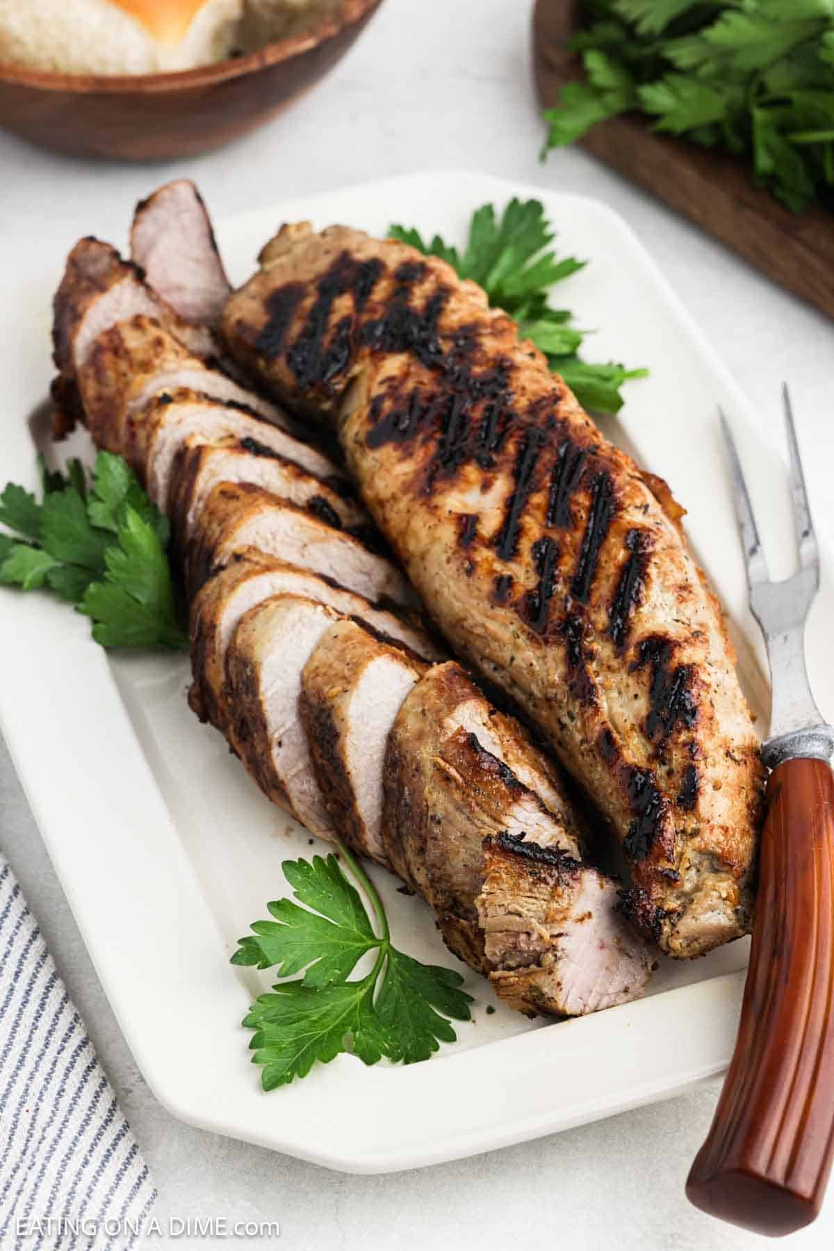 Cooked pork tenderloin sliced and placed on a white platter with fresh herbs on the side.