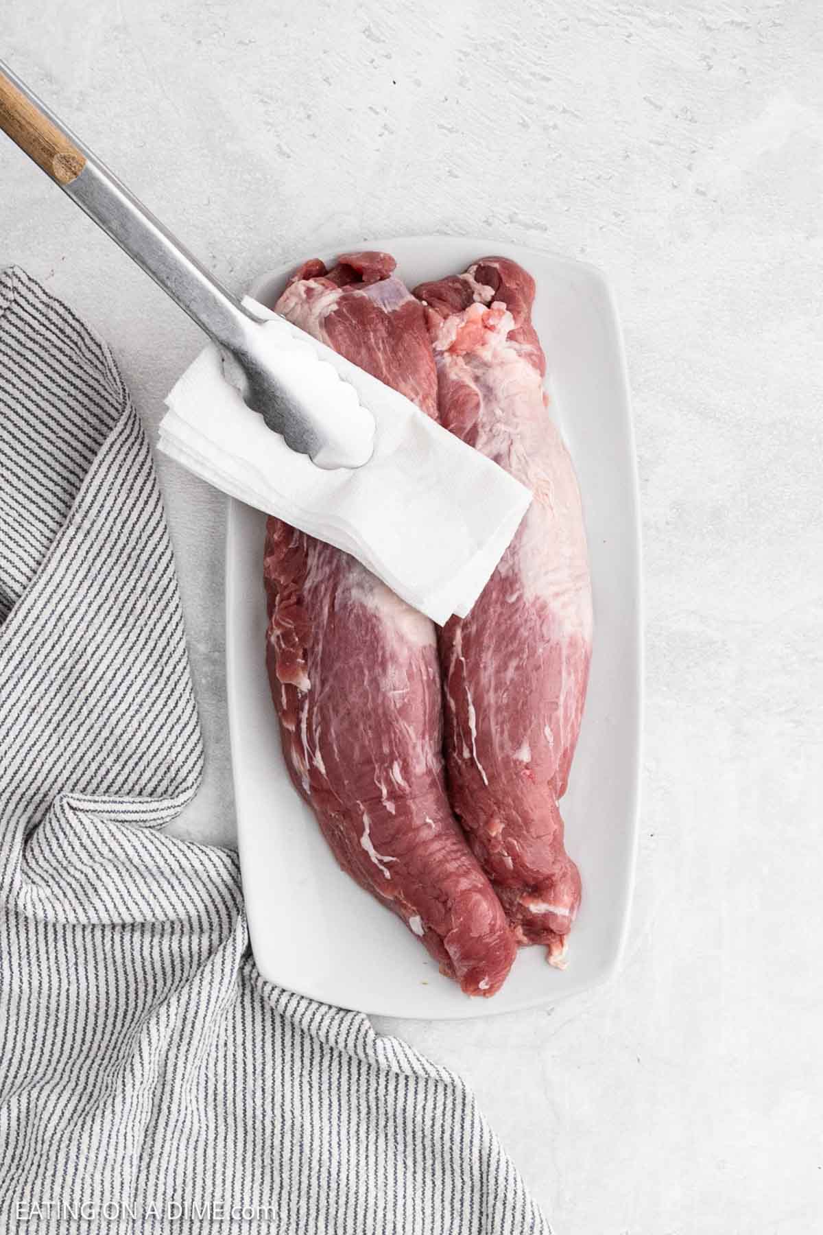 Raw pork tenderloin on a platter with a paper towel in between tongs patting the pork dry.