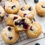 Golden blueberry muffins from an Easy Blueberry Muffin Recipe rest on a cooling rack, topped with sugar. One muffin is cut in half, revealing a moist, fluffy interior filled with juicy blueberries. Whole blueberries are scattered nearby.