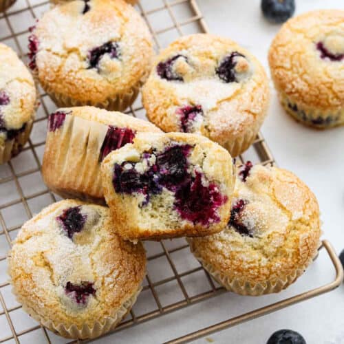 Golden blueberry muffins from an Easy Blueberry Muffin Recipe rest on a cooling rack, topped with sugar. One muffin is cut in half, revealing a moist, fluffy interior filled with juicy blueberries. Whole blueberries are scattered nearby.