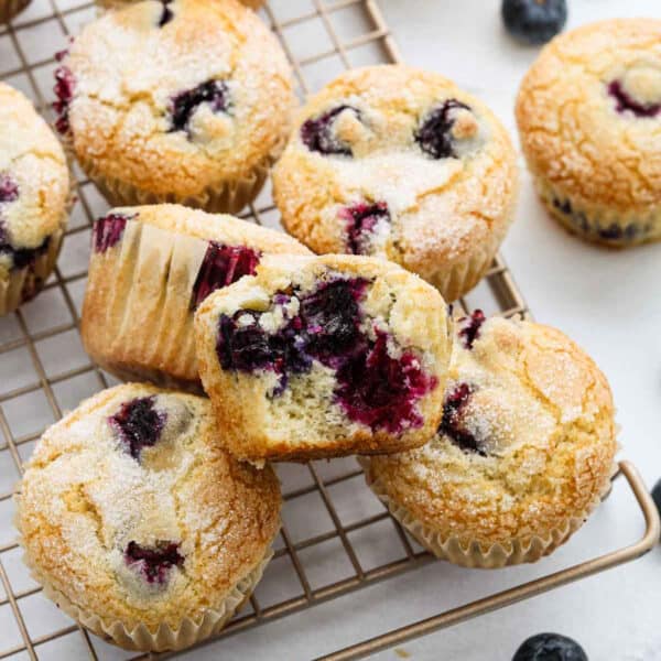 Golden blueberry muffins from an Easy Blueberry Muffin Recipe rest on a cooling rack, topped with sugar. One muffin is cut in half, revealing a moist, fluffy interior filled with juicy blueberries. Whole blueberries are scattered nearby.