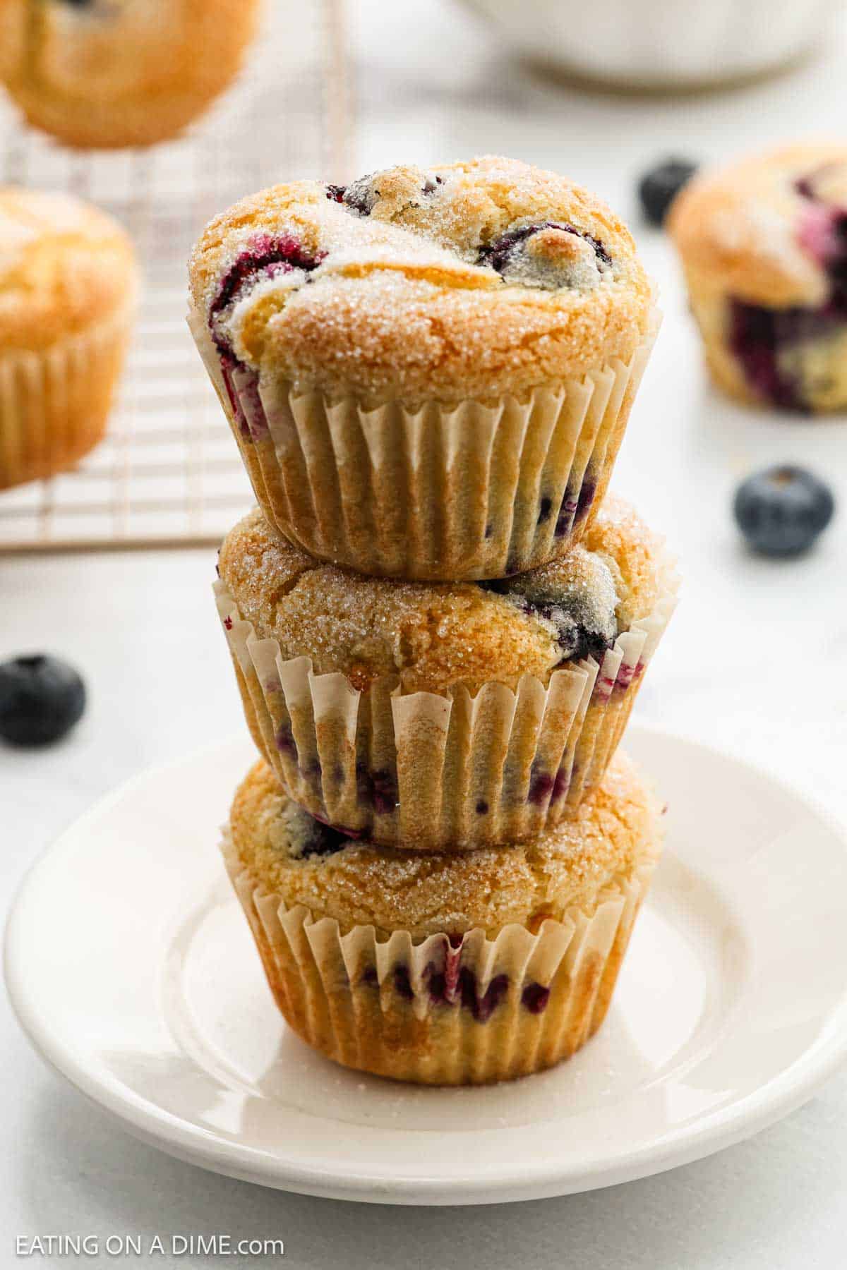 Three sugar-dusted blueberry muffins stacked on a white plate, made using an Easy Blueberry Muffin Recipe, with fresh blueberries and more muffins visible in the background.