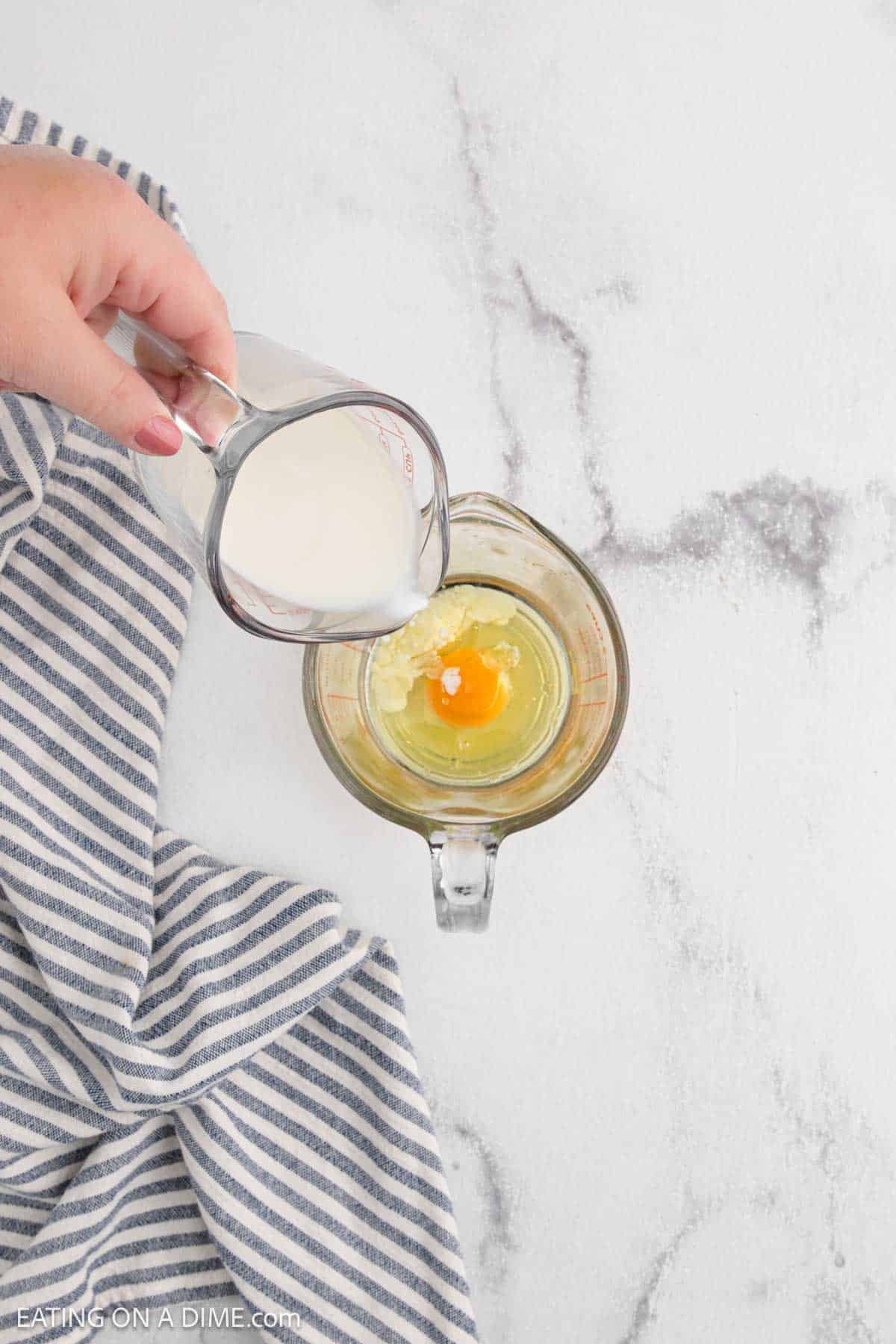 A hand pours milk from a measuring cup into a glass measuring cup containing a cracked egg, next to a blue and white striped clothโan essential step in this Easy Blueberry Muffin Recipe.