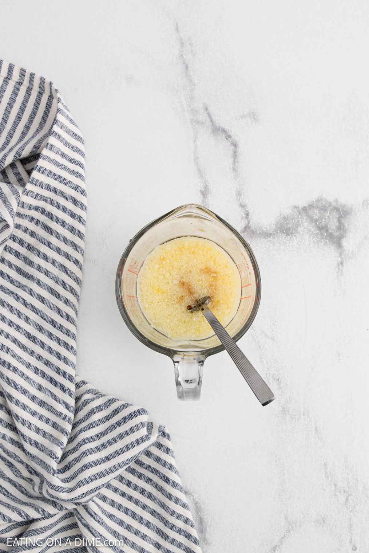 A glass measuring cup filled with a yellowish liquid and bubbles, with a metal spoon inside, sits on a white marble surface next to a blue and white striped kitchen towelโperfect for mixing your Easy Blueberry Muffin Recipe.