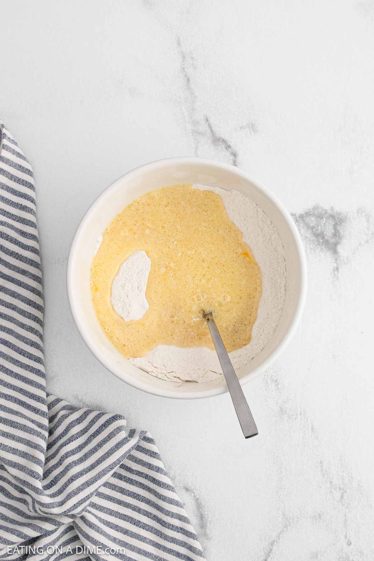 A mixing bowl with flour and a yellow liquid mixture, partially combined with a metal spoonโjust the start of an Easy Blueberry Muffin Recipe. A blue and white striped kitchen towel sits beside the bowl on a light-colored surface.