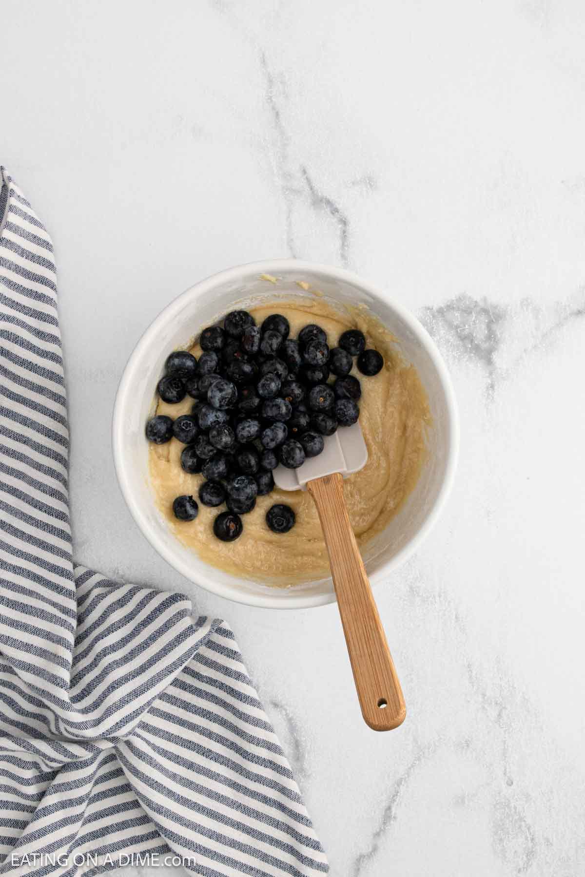 A white bowl filled with blueberry muffin batter and fresh blueberries, ready to be mixed for this Easy Blueberry Muffin Recipe. The bowl rests on a white marble surface beside a blue-and-white striped cloth.