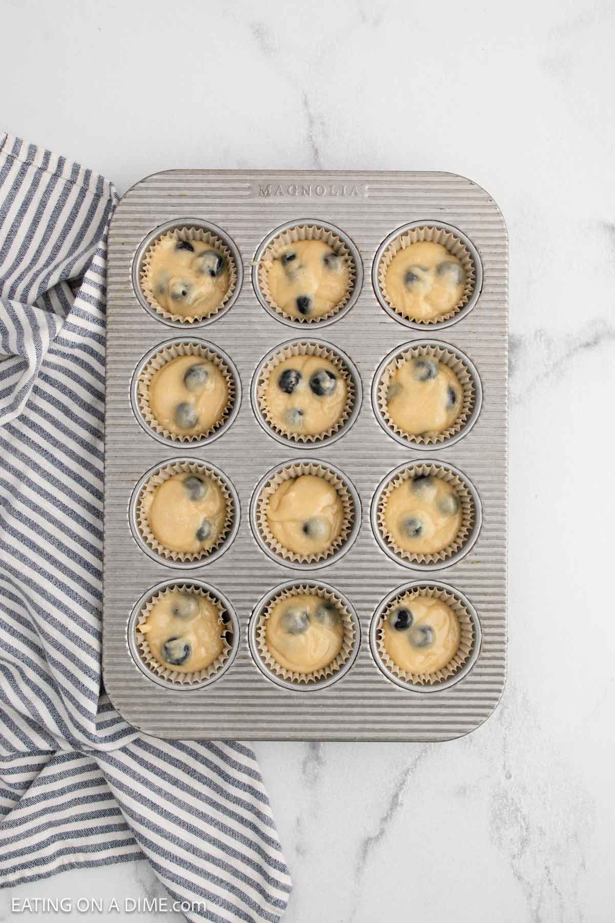 A muffin tin filled with cupcake liners containing unbaked batter from an easy blueberry muffin recipe sits on a white marble surface next to a striped kitchen towel.