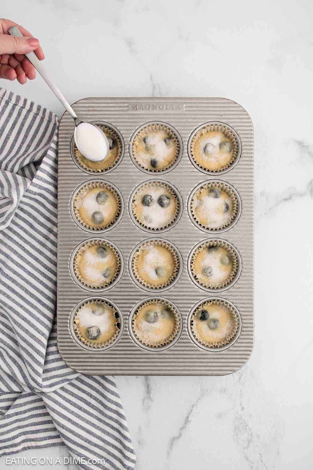 A hand sprinkles sugar with a spoon over unbaked batter from an Easy Blueberry Muffin Recipe in a 12-cup muffin tin lined with paper liners, on a marble countertop next to a striped kitchen towel.