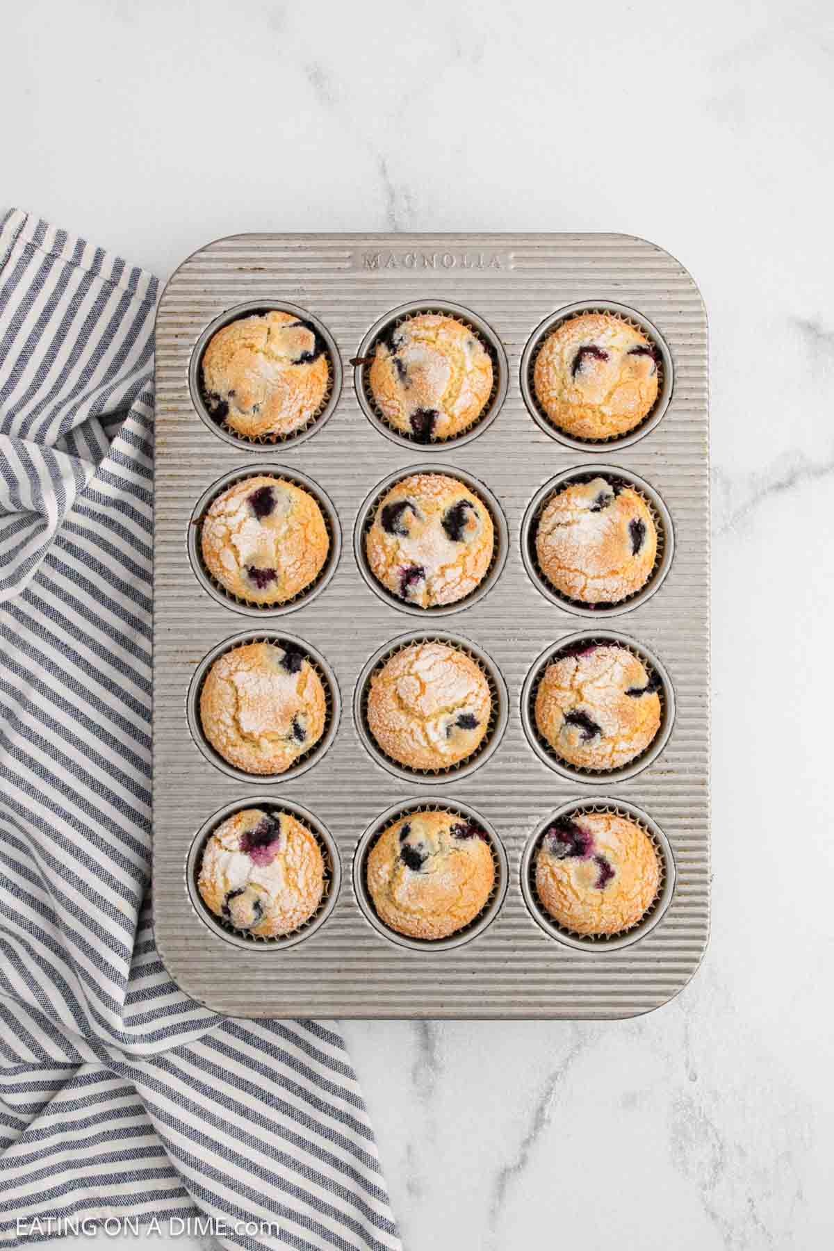 A muffin tin filled with twelve golden-brown treats from an Easy Blueberry Muffin Recipe, some with juicy blueberries on top, sits on a marble surface beside a striped gray and white kitchen towel.