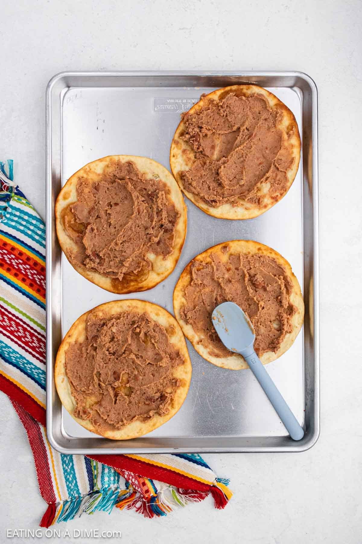 Spreading refried beans over the crispy tortillas on a baking sheet.