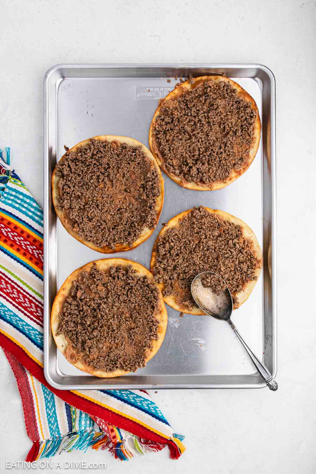Cooked ground beef on refried beans tortillas on a baking sheet.