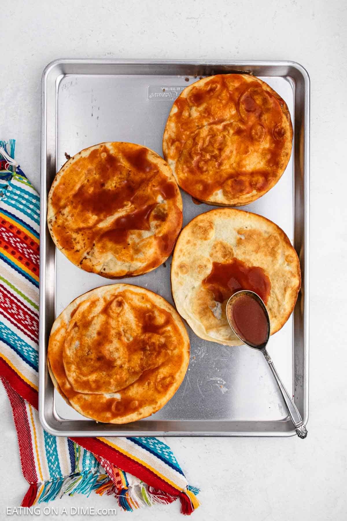 Spreading red enchilada sauce over the crispy tortillas on a baking sheet.