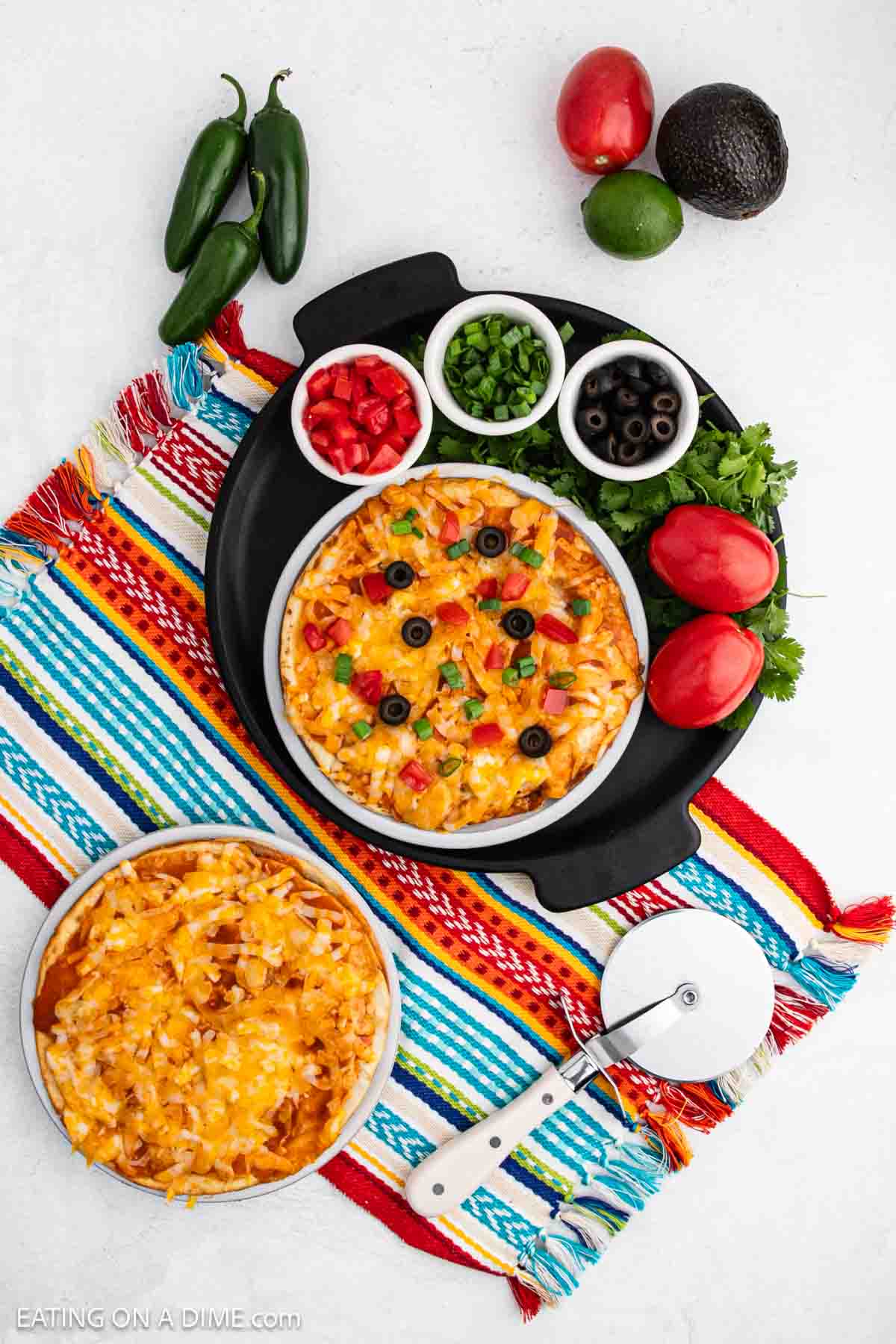 Mexican pizza topped with melted cheese, black olives, and tomatoes with small bowls of toppings nearby.