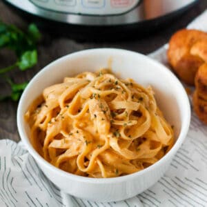 A bowl of creamy pasta topped with herbs, placed on a textured table. In the background, an Instant Pot is visible, and there are small rolls beside the bowl. The dish represents Instant Pot Buffalo Chicken Pasta.
