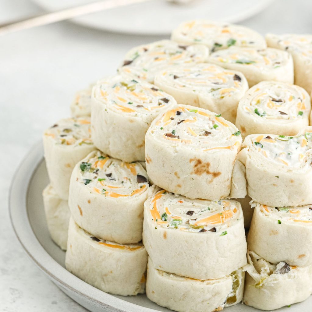 A plate stacked with neatly arranged cream cheese tortilla roll ups, filled with shredded cheese and chopped vegetables.