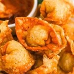 Close-up of several crispy, golden-brown fried wontons from our delicious Fried Wonton Recipe, with a small bowl of dipping sauce in the background. The wontons have a bubbly texture and are arranged in a visually appealing pile.