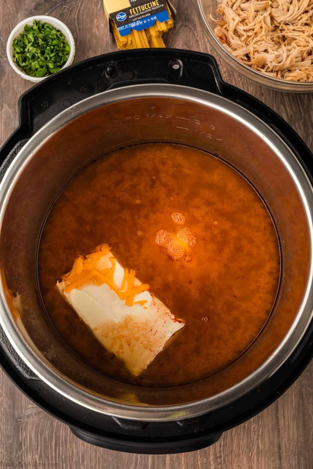 An Instant Pot Buffalo Chicken Pasta scene features broth, a block of cream cheese, and shredded cheddar inside. Nearby are bowls of chopped herbs, shredded chicken, and uncooked fettuccine pasta, all atop a wooden surface.