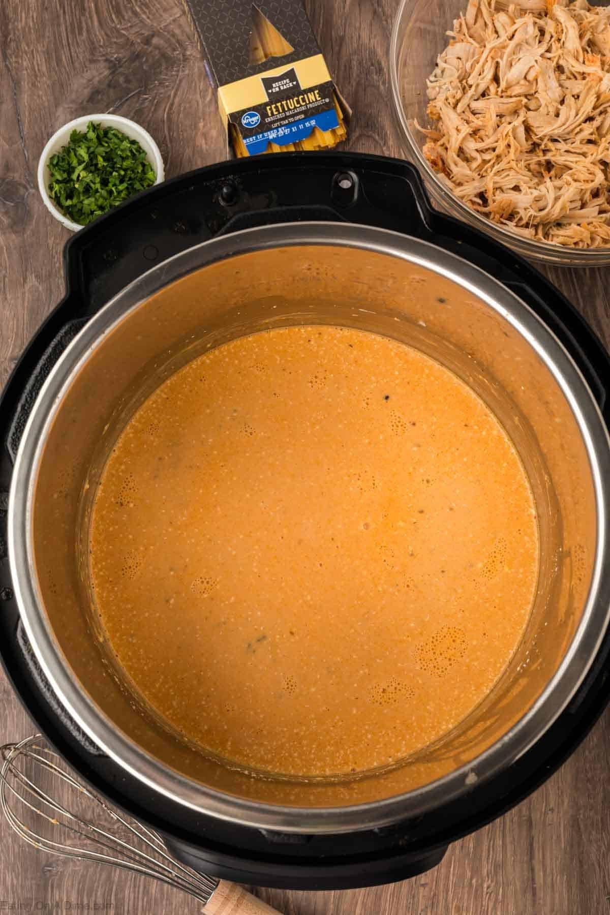 An Instant Pot Buffalo Chicken Pasta sits on a wooden surface, filled with creamy orange sauce and surrounded by a bowl of shredded chicken, fettuccine pasta, and a small bowl of chopped green herbs.