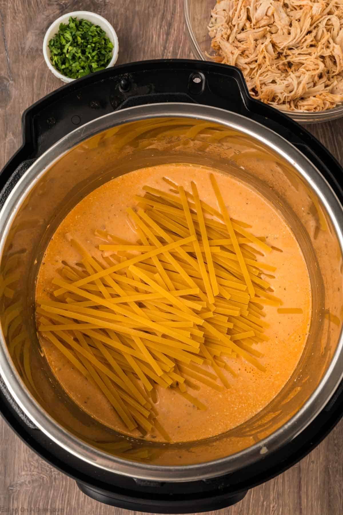 An Instant Pot filled with creamy orange sauce and uncooked noodles, perfect for Instant Pot Buffalo Chicken Pasta. In the background, a bowl of shredded chicken and a small bowl of chopped herbs sit on a wooden surface.