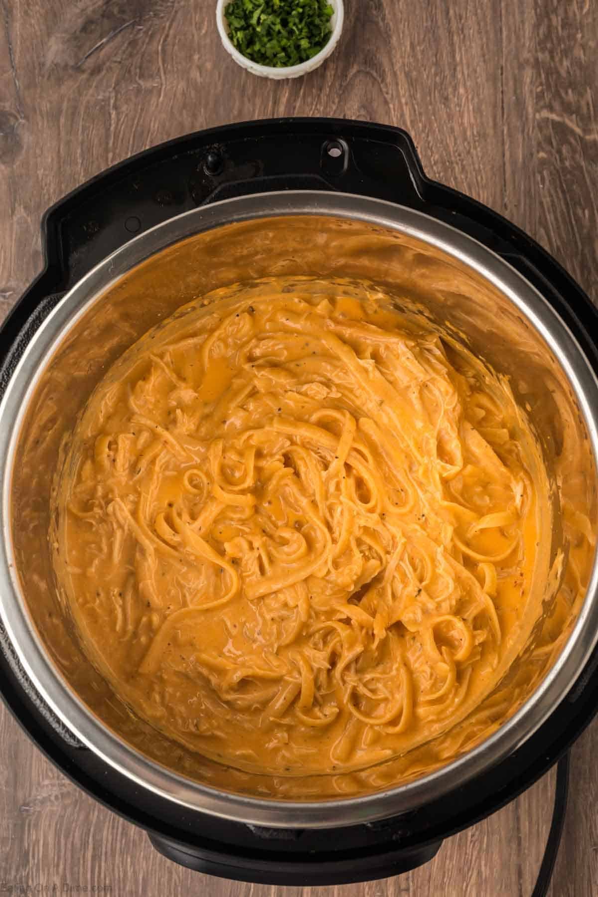 A pot filled with creamy Instant Pot Buffalo Chicken Pasta in a thick orange sauce sits on a wooden table. A small bowl of chopped green herbs is visible in the background.