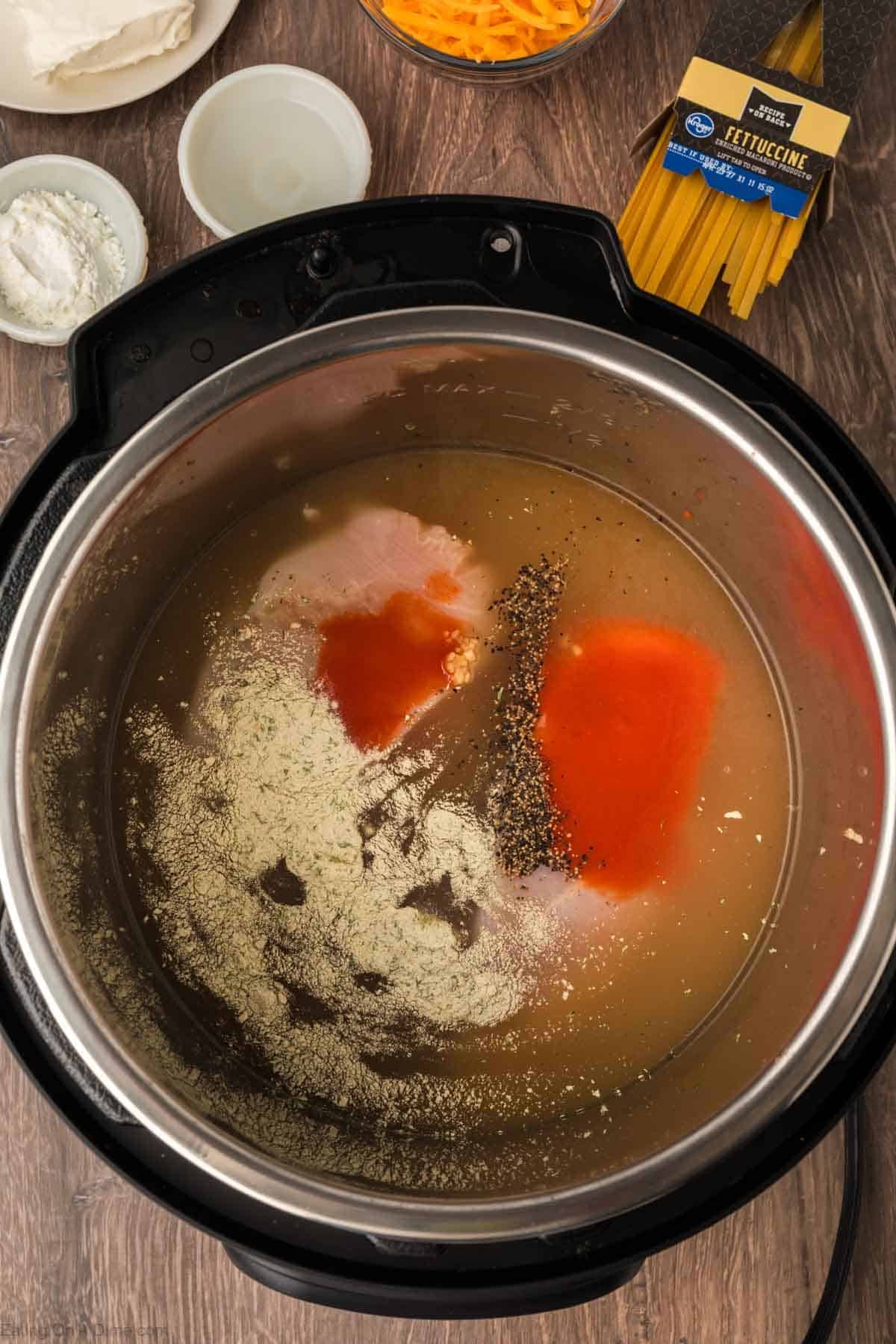 An Instant Pot Buffalo Chicken Pasta is prepped with uncooked noodles, broth, hot sauce, pepper, and seasonings. Nearby bowls hold shredded cheese, cream cheese, and cornstarch on a wooden surface.
