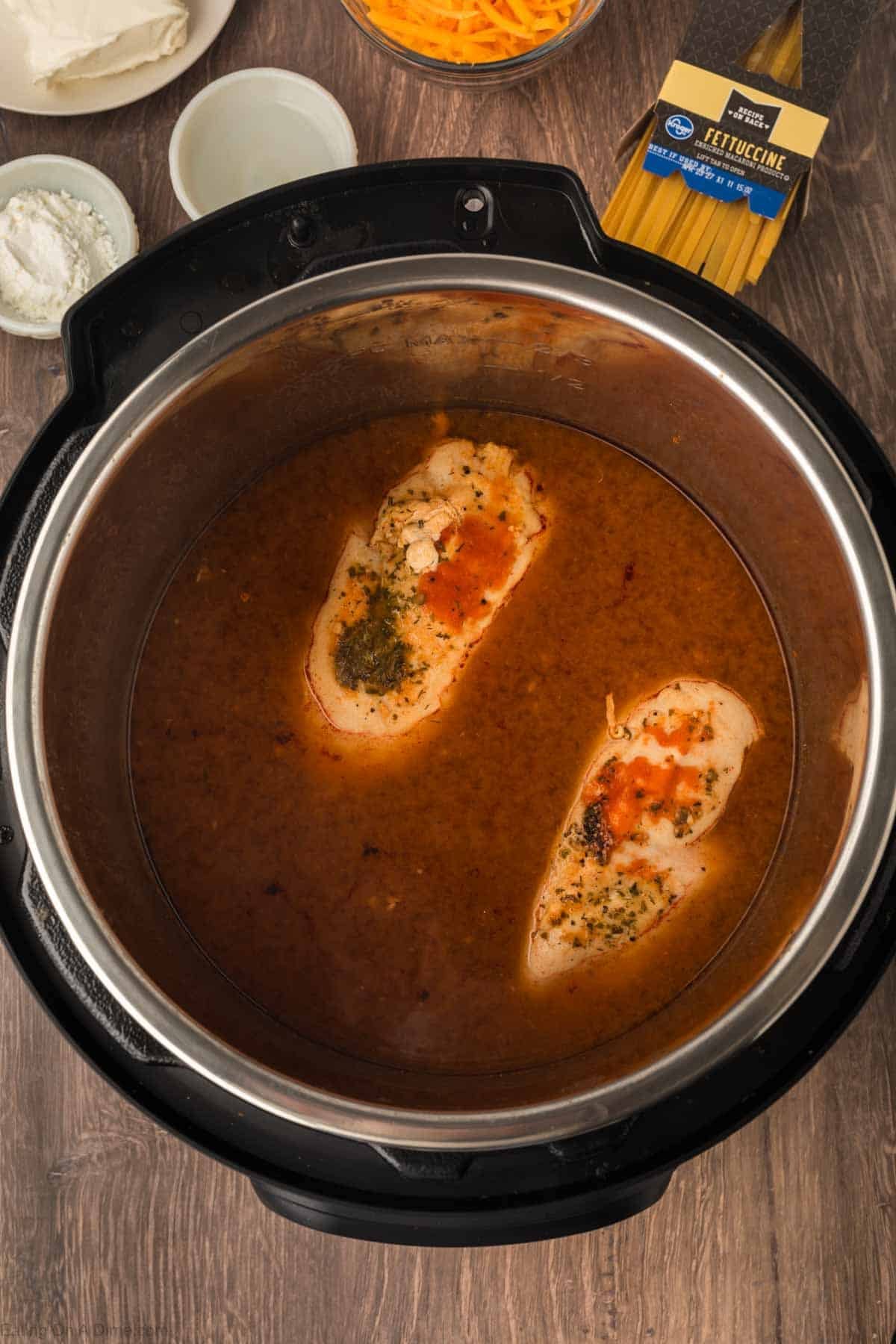 Two seasoned chicken breasts simmer in flavorful liquid inside an Instant Pot, surrounded by dried fettuccine, shredded cheese, cream cheese, and flour—perfect for making creamy Instant Pot Buffalo Chicken Pasta.