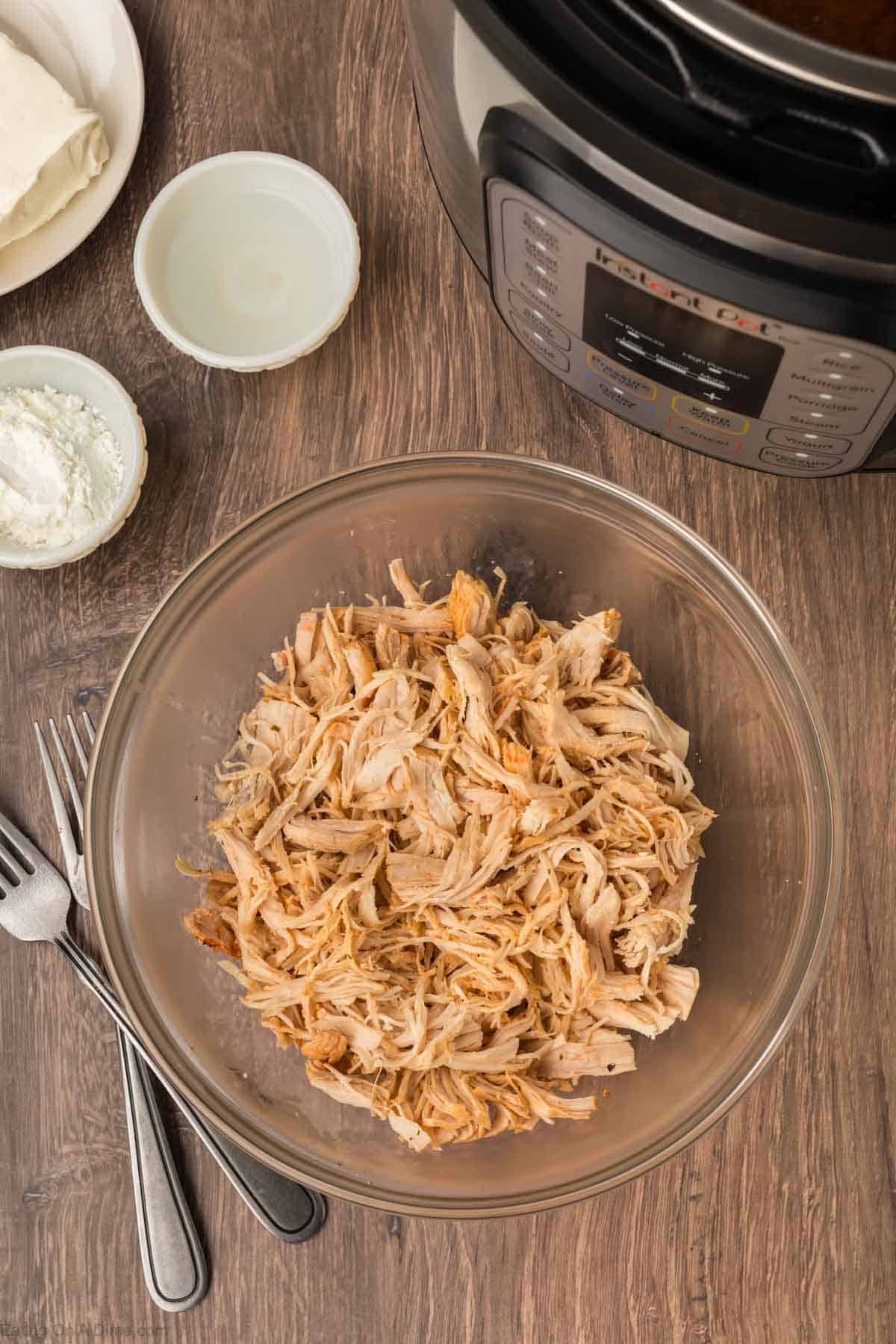 A glass bowl filled with shredded chicken sits on a wooden surface next to an Instant Pot, two forks, and small bowls of cream cheese and cornstarch—perfect ingredients for making Instant Pot Buffalo Chicken Pasta.